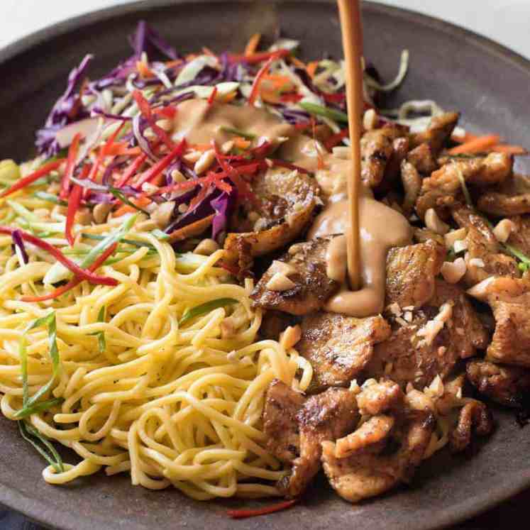 Peanut dressing being poured over Satay Chicken Noodle Salad in a brown bowl.